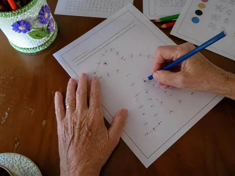 Mãos de uma senhora fazendo uma atividade no papel, com lápis de cor na mesa